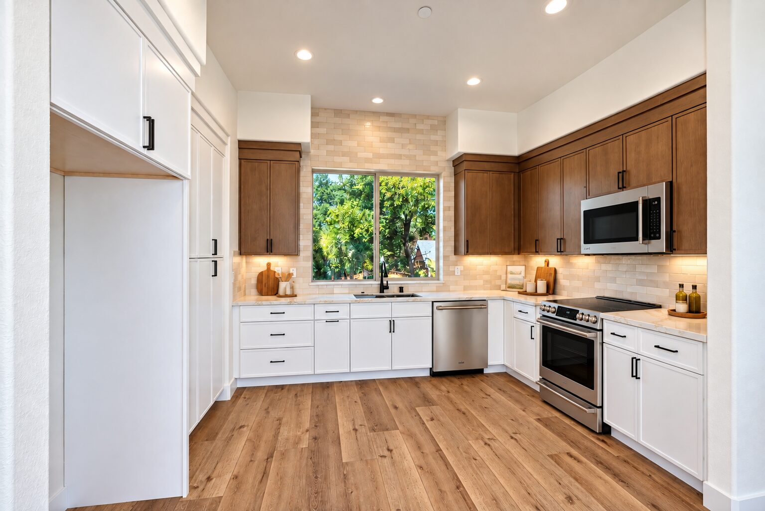 Bright, modern kitchen with white lower cabinets, warm wood upper cabinets, black hardware, stainless-steel appliances, and wide-plank wood flooring. A large window above the sink overlooks lush green trees, while recessed and under-cabinet lighting illuminate the space.